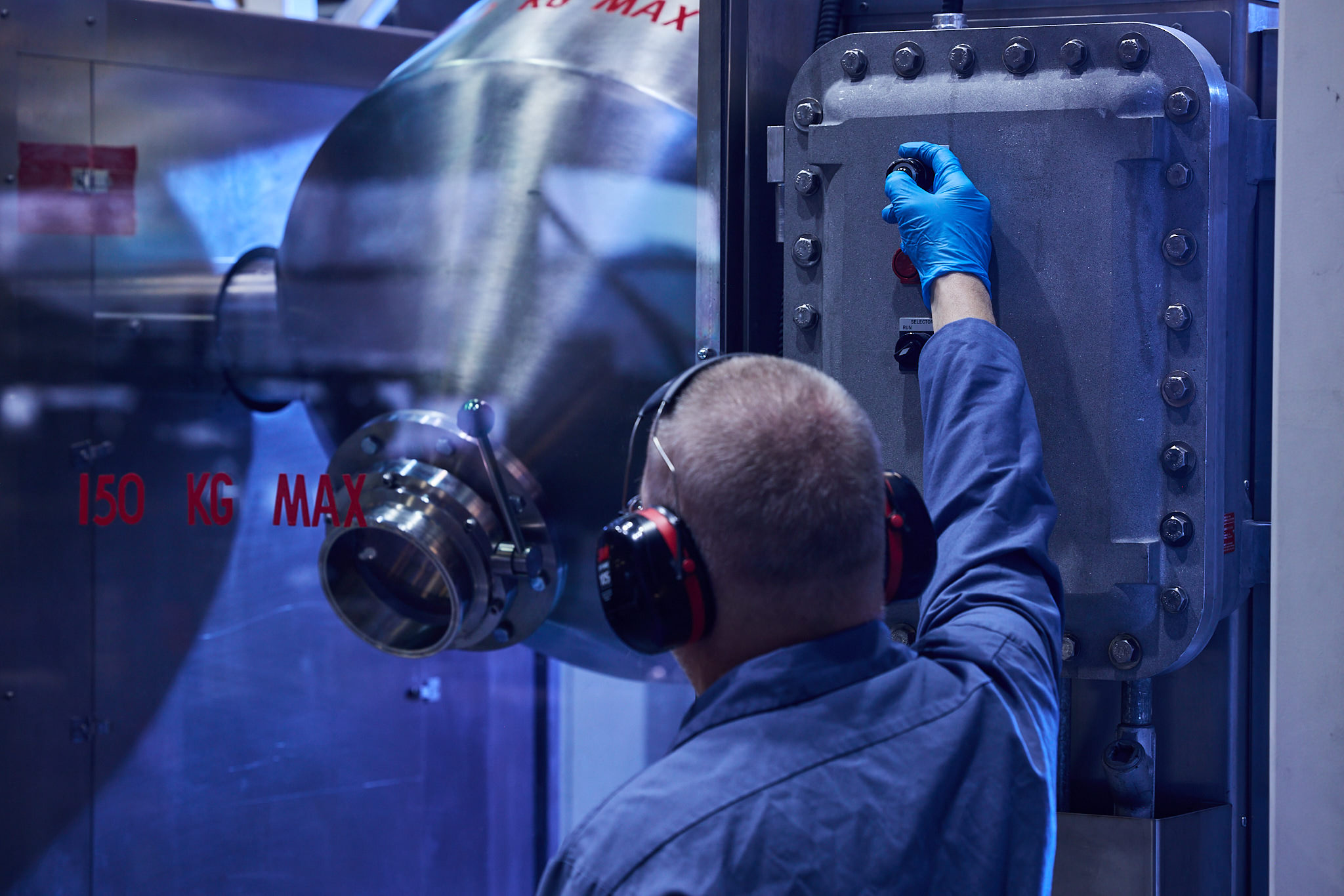 Man in blue workwear and ear protection operates industrial machinery, reaching for control panel with gloved hand, near large metal equipment with safety labels indicating weight capacity.