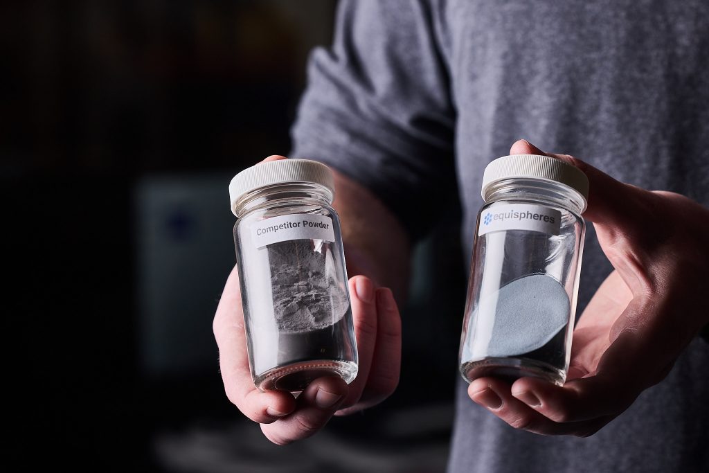 Two hands are holding glass bottles filled with aluminum powder, with the one labelled Equispheres being more densely packed. 